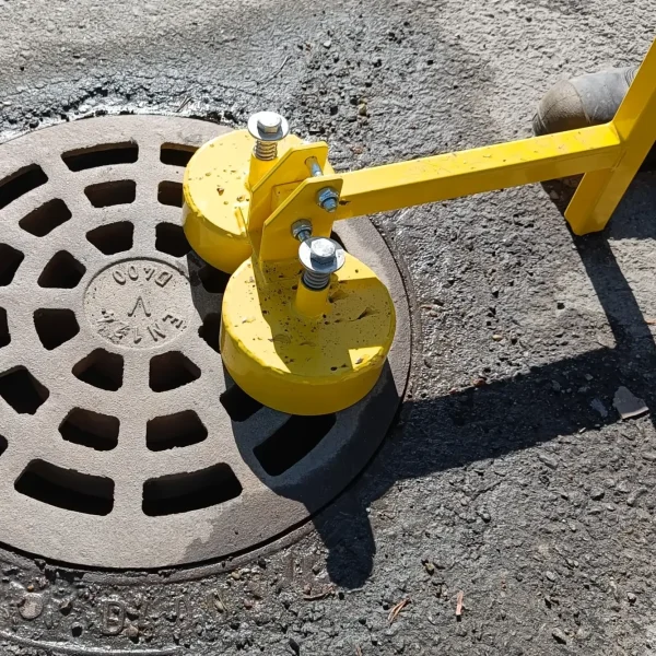 Lähikuva keltaisesta KM 1200 kaivonkansimagneettista, joka on kiinnittynyt valurautaiseen sadevesikaivon kanteen asfaltilla.