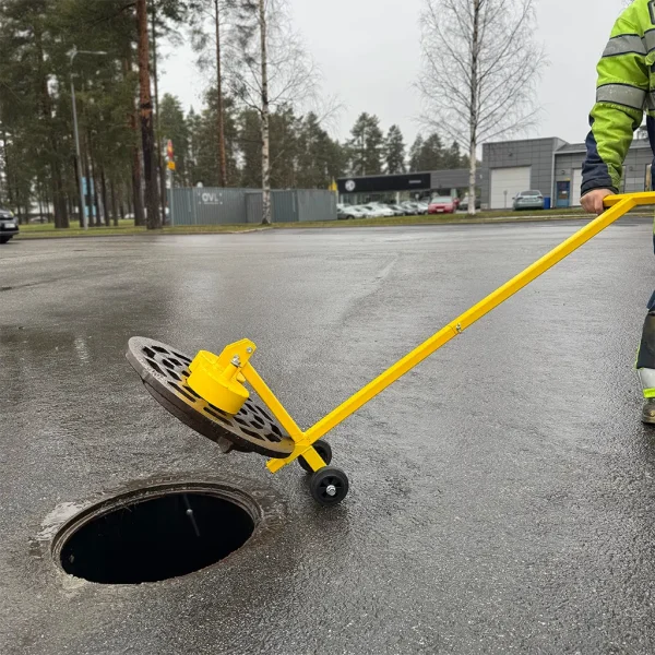 Työntekijä on avannus valurautaisen kaivonkannen KM1200 kansimagneetilla, jossa on lisävarusteena asennettuna pyörät. Pyörät tekevät työkalun liikuttelusta kevyempää.