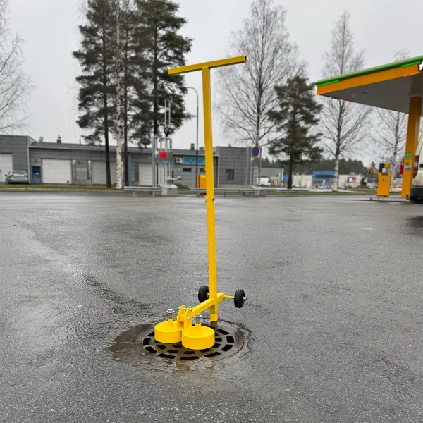 Keltainen KM1200 kahdella kestomagneetilla ja pyörillä varustettu kaivonkannen avaustyökalu seisoo asfaltilla liikenneaseman pihassa.