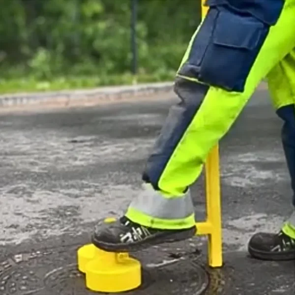 Työasuinen henkilö käyttää keltaista kaivonkannen avaustyökalua ja aktivoi kestomagneetit painamalla jalalla laitteen jousimekanismin alas, kun laite on kohdistettu rautakannelle.